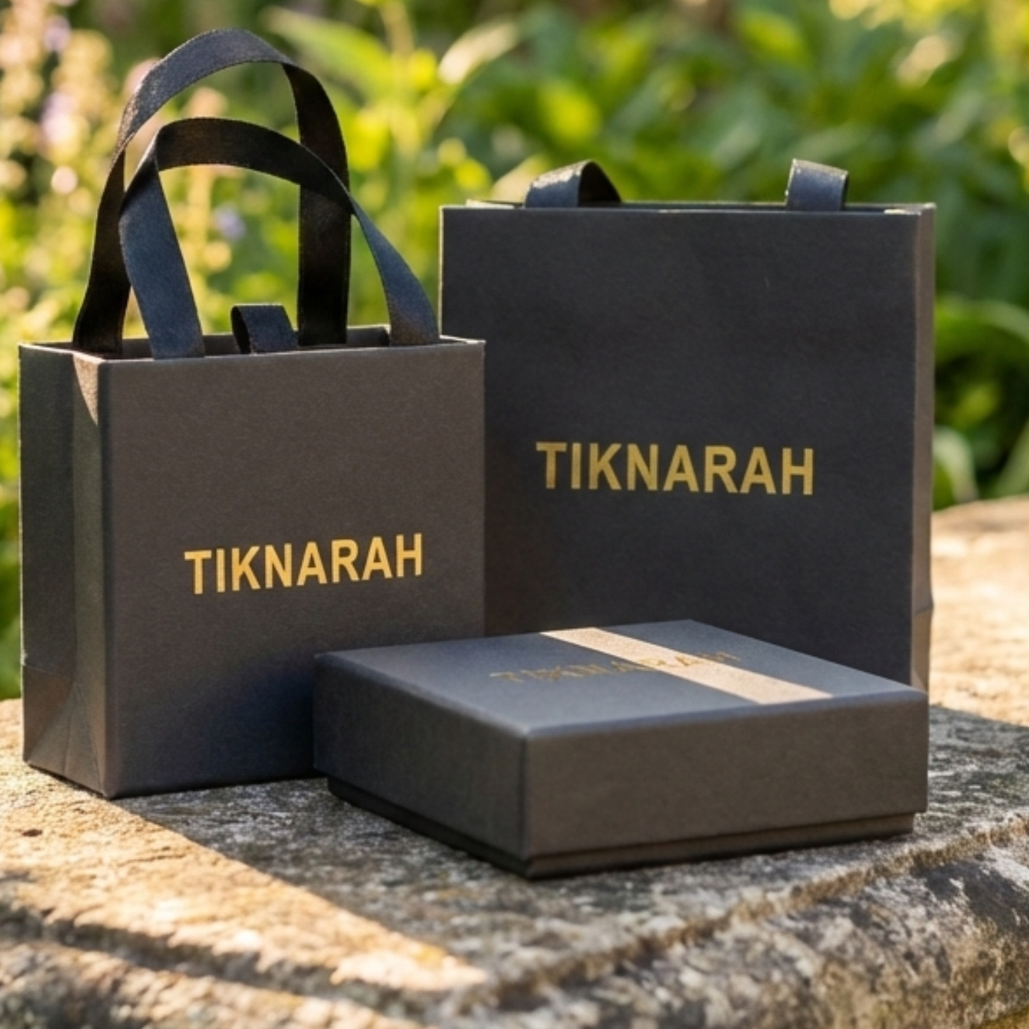 Black gift bags and box with 'TIKNARAH' branding on a stone surface with greenery in the background.