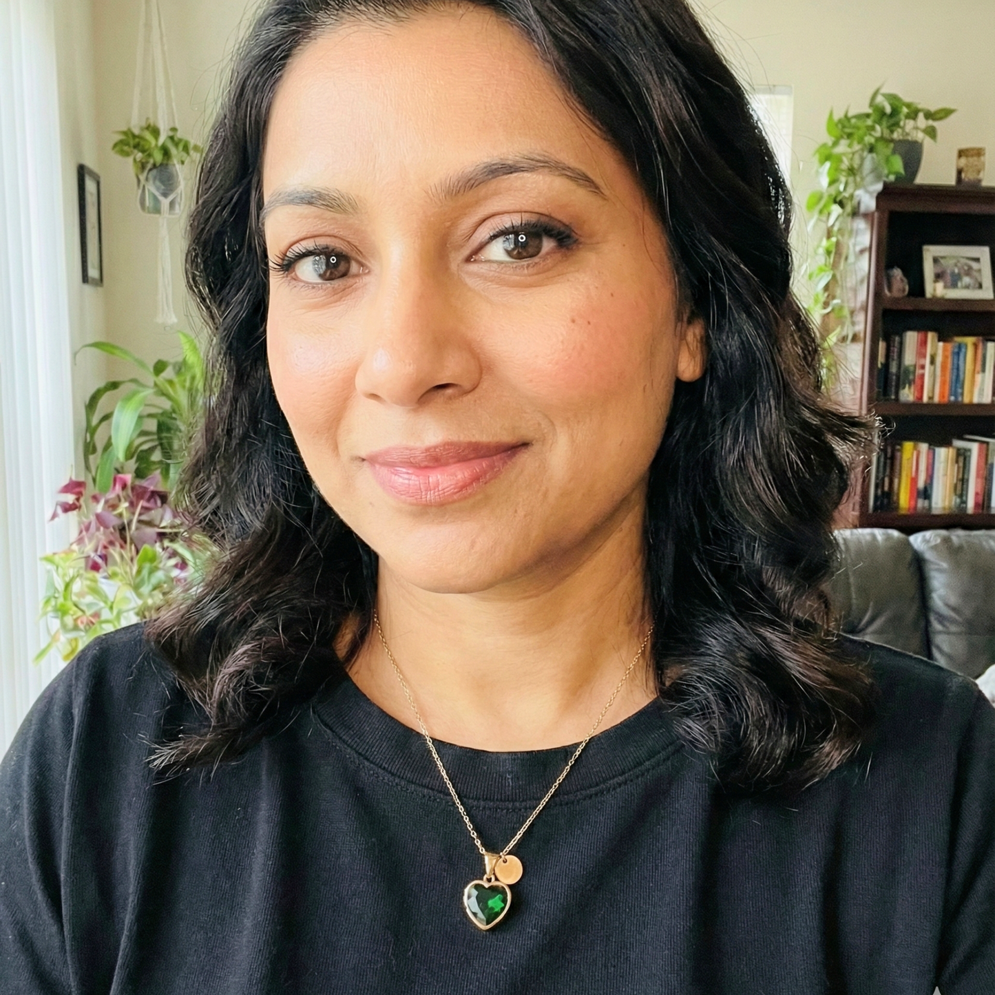 Woman wearing a black shirt with a heart-shaped pendant necklace in a home setting. Tiknarah, emerald heart pendant, rose gold lariat, green stone necklace, romantic gift for her, Y-drop jewelry, luxury fashion necklace, Shopify jewelry store, minimalist emerald jewelry.